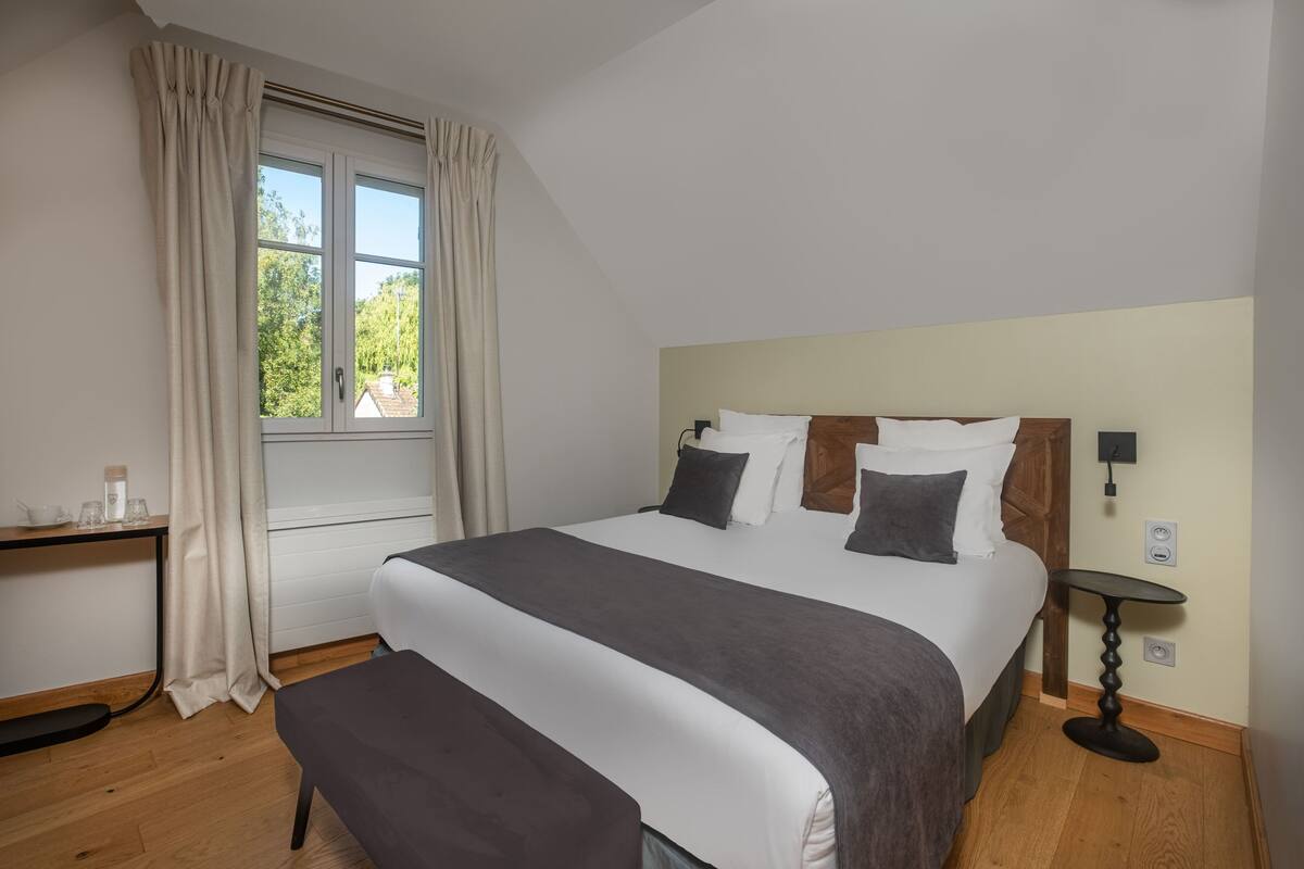 Ein ordentlich eingerichtetes Schlafzimmer mit einem großen Bett, einem Nachttisch, einem Fenster mit Vorhängen und Blick ins Grüne.