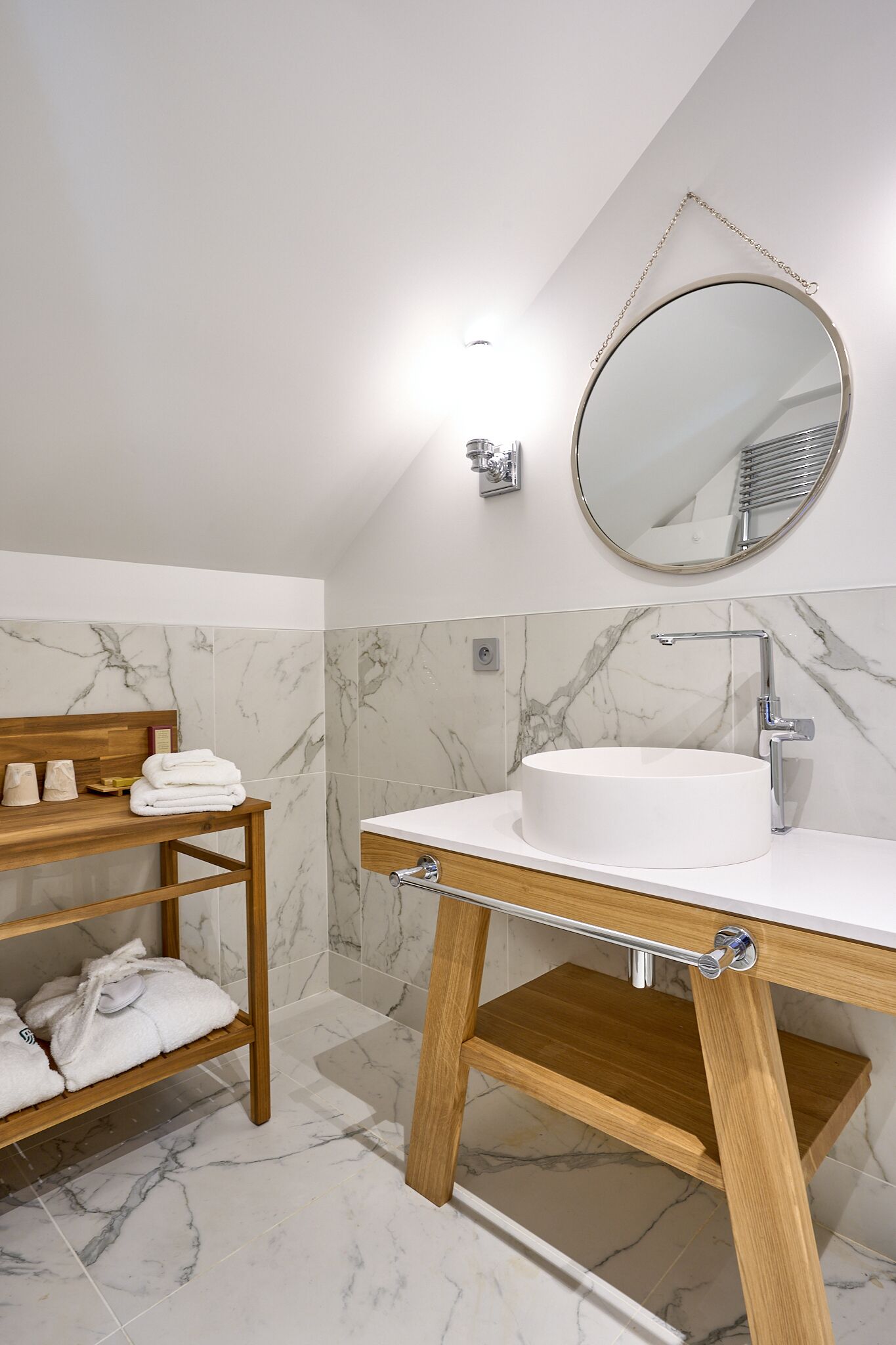 Un baño con pared revestida de mármol, tocador de madera con lavamanos, espejo redondo y estante con toallas.