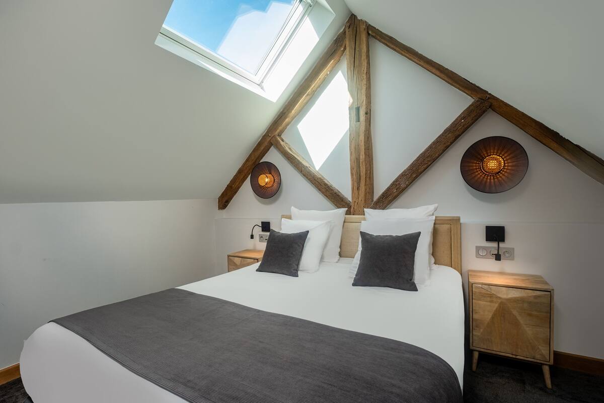 Ein Schlafzimmer mit einem großen Bett, Holzbalken, einem Oberlicht, zwei Nachttischen und einer wandmontierten Leuchte.