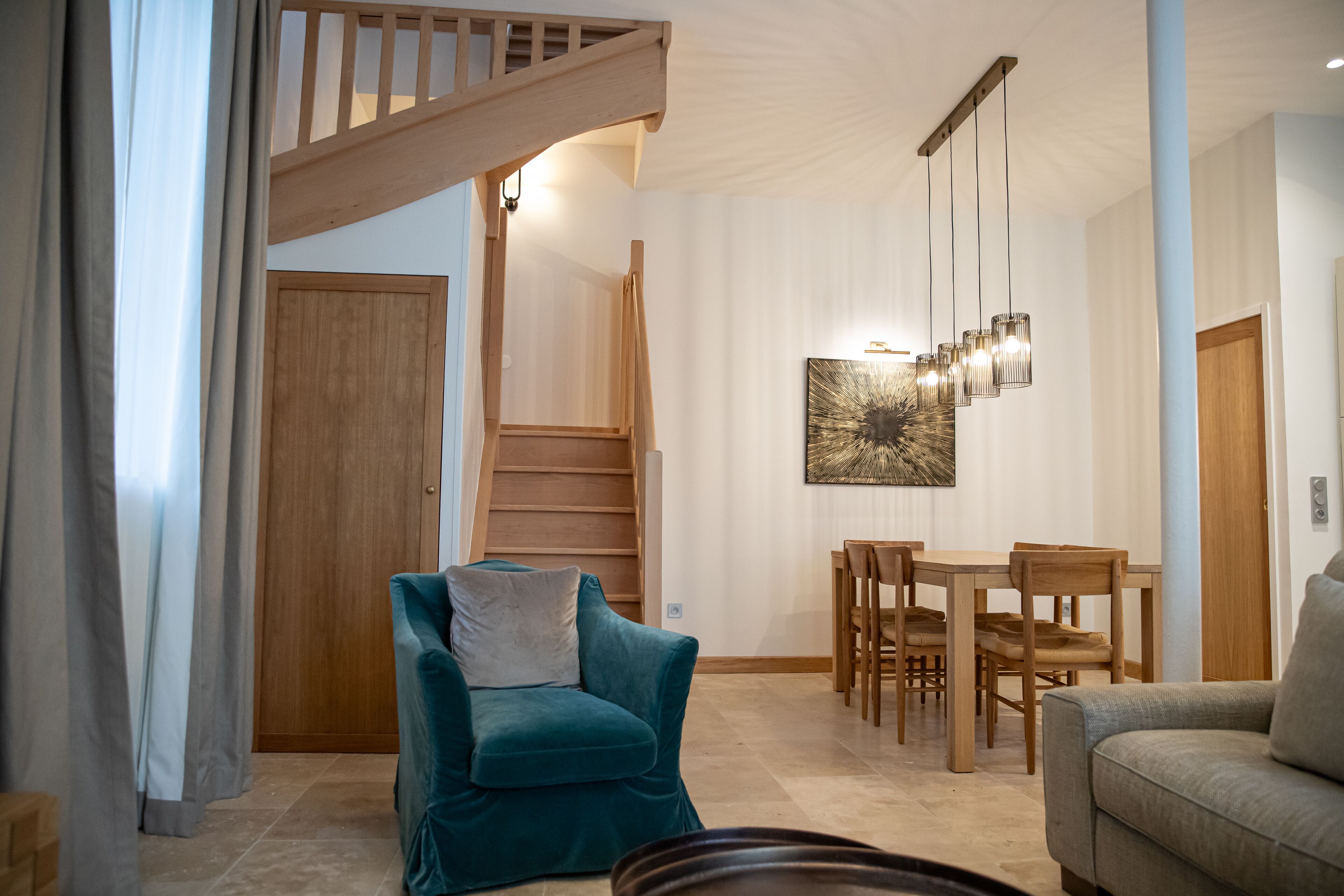A spacious living room with a teal armchair, a gray sofa, a wooden dining table with chairs, and a staircase leading to an upper level.