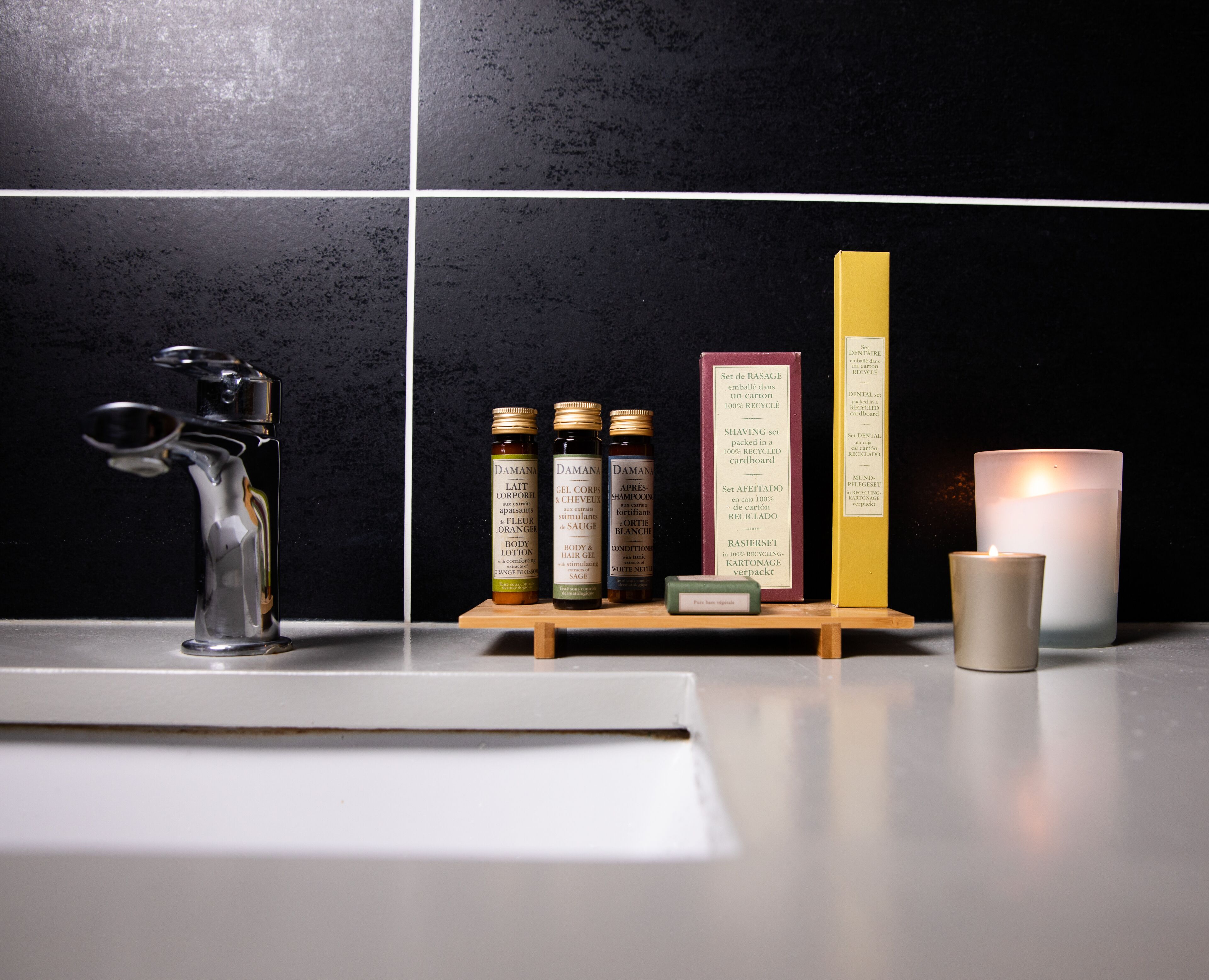 A bathroom counter with a faucet, three bottles of shampoo, a box of razors, and a lit candle.