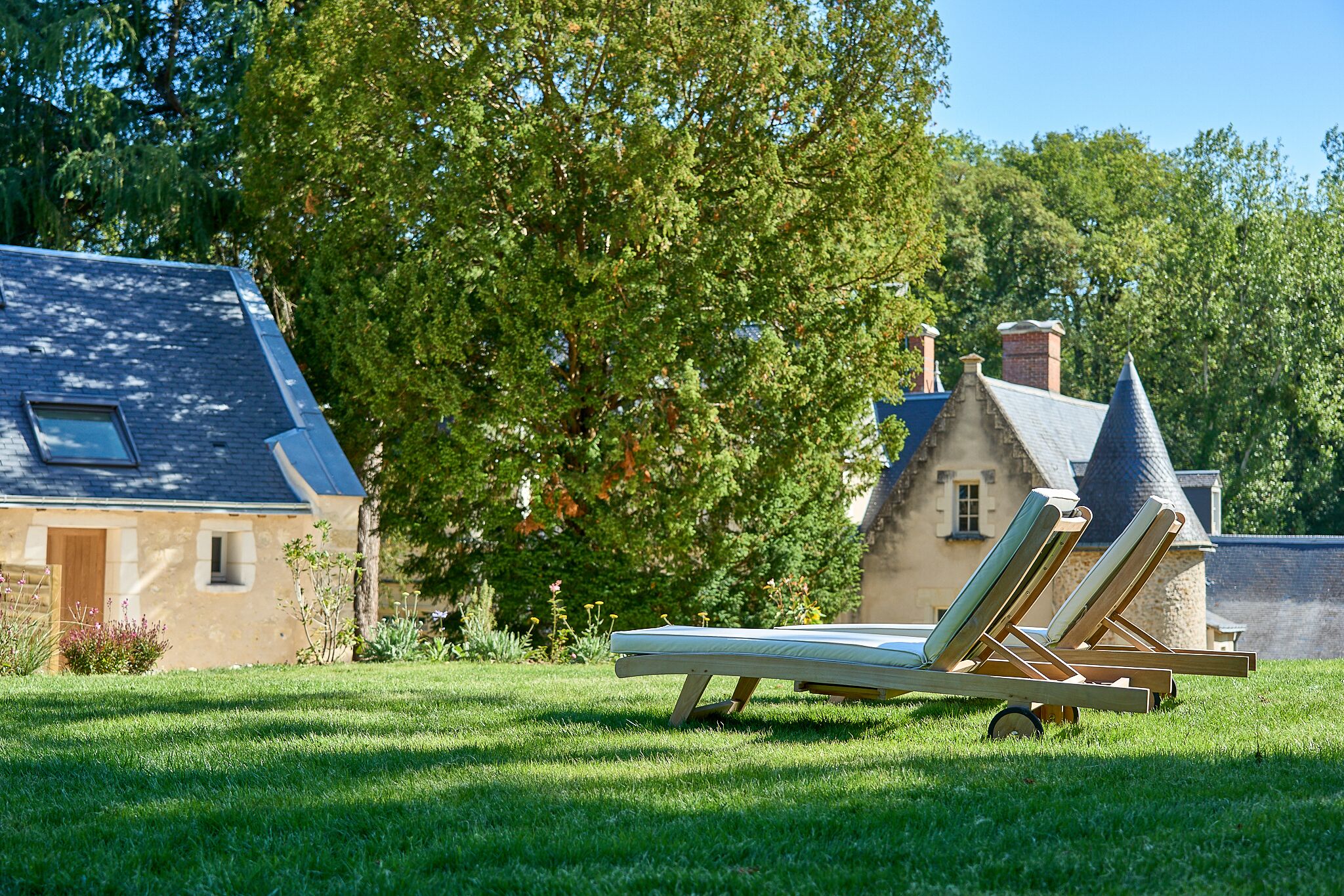 A grassy area with two lounge chairs and a mattress, a stone building with a sloped roof, and a large tree in the background.