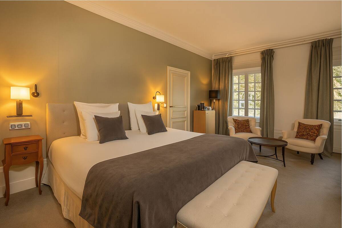 Ein Hotelzimmer mit einem großen Bett, Nachttischlampen, einem Sessel, einem kleinen Tisch und einem Fenster mit Vorhängen.