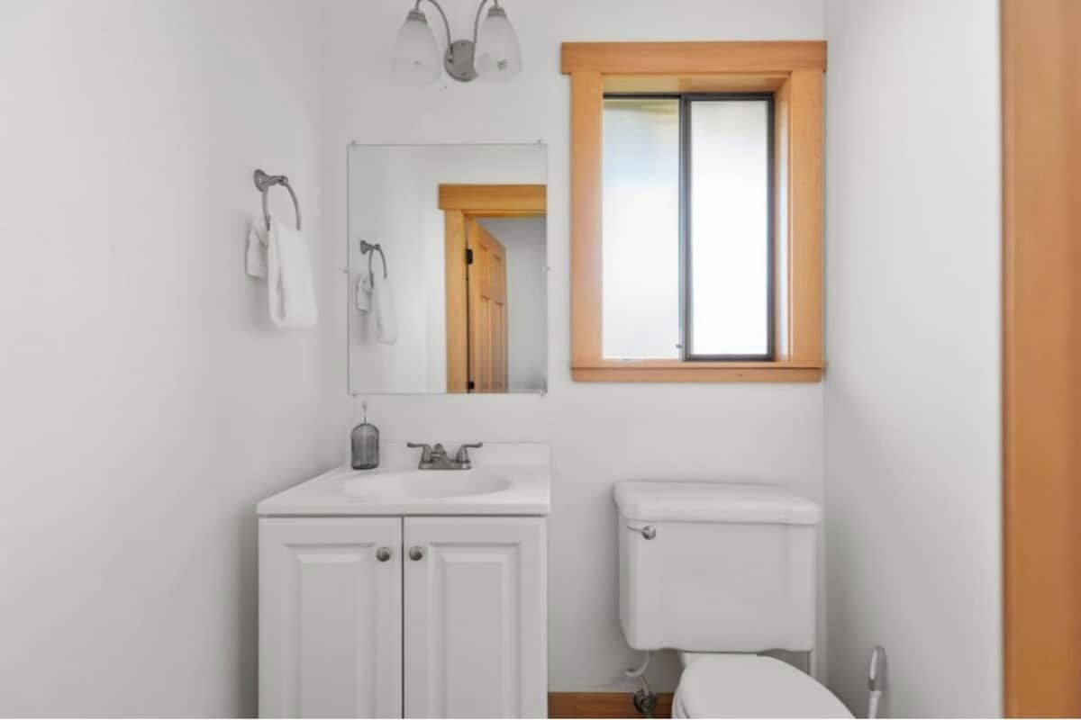 Ein kleines, sauberes Badezimmer mit Waschbecken, Spiegel und Toilette in Weiß. Ein Fenster mit Holzrahmen und ein Handtuch an der Wand.
