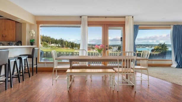 A spacious dining area with a wooden table and chairs, large windows with a view of a lake and greenery, and a kitchen with wooden cabinets.
