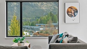 A living room with a large window offering a view of a mountain and buildings. There is a framed picture on the wall and a potted plant on a side table. - Luxury Penthouse with Mountain Views in Queenstown (Queenstown)