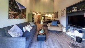 Una sala de estar con sofá, sillón, mesa de centro y televisor. Hay un cuadro en la pared y una cocina al fondo.