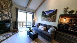 Un salón acogedor con chimenea de piedra, un sofá oscuro con cojines, una mesa de centro de madera y un cuadro grande con paisaje de montaña.