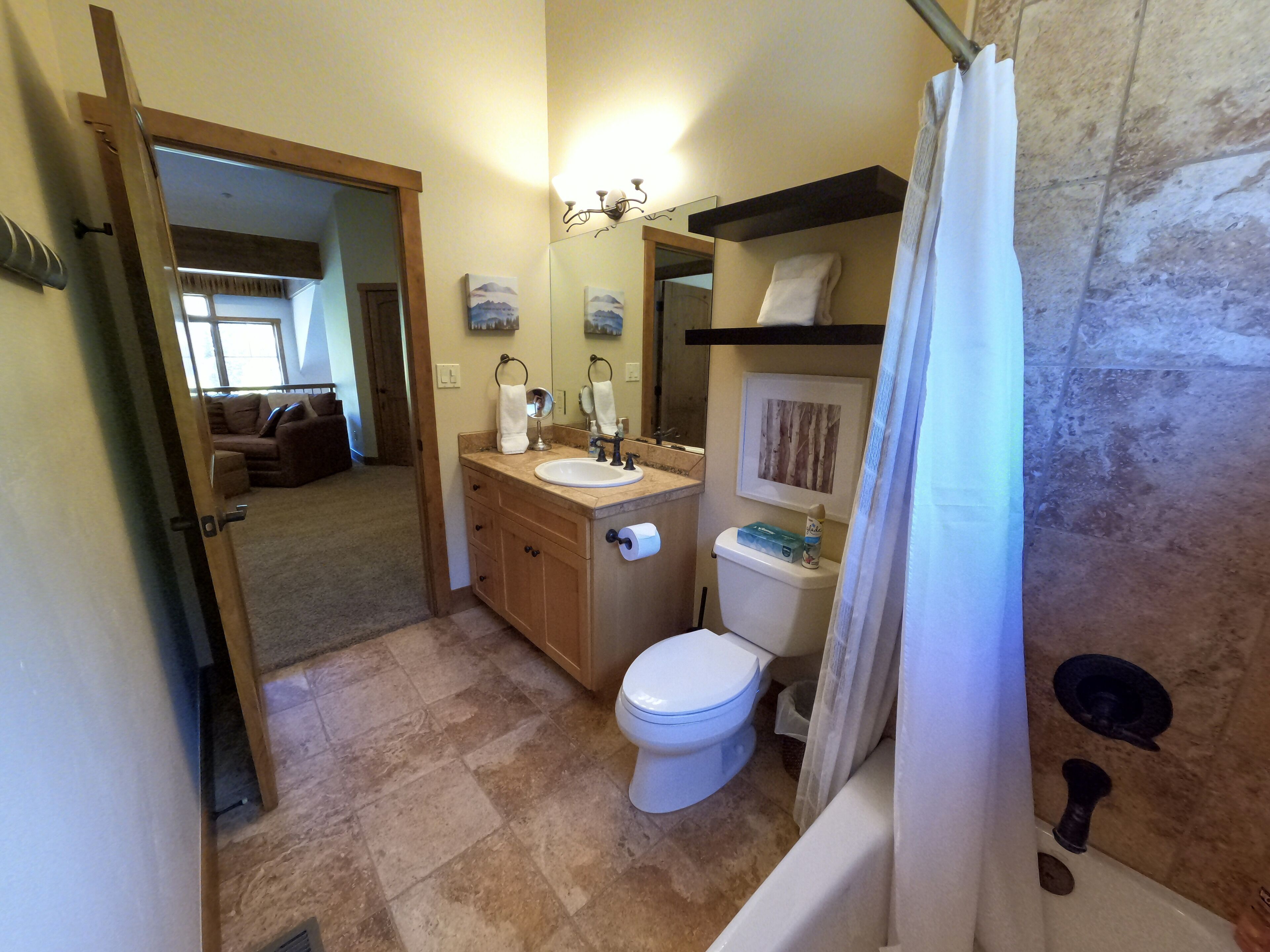 Un baño con inodoro, lavabo y ducha. Hay un espejo sobre el lavabo y un perchero para toallas. El baño tiene suelo de baldosas y una cortina de ducha.