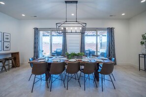 Un coin repas avec une grande table dressée pour six personnes, entourée de chaises, et un lustre moderne au-dessus.