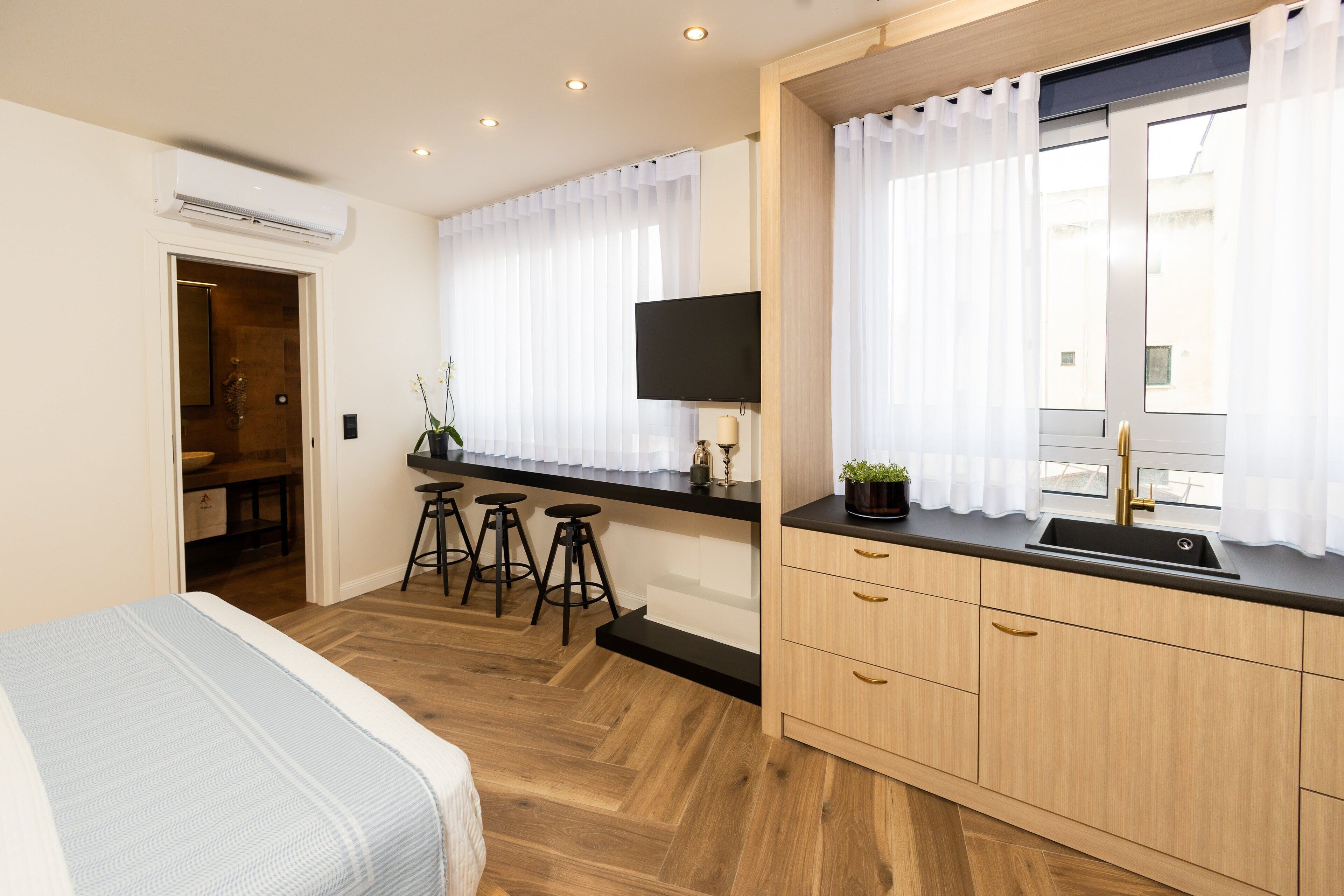 A modern bedroom with a bed, a desk with a TV, and a kitchen area with a sink and stools.