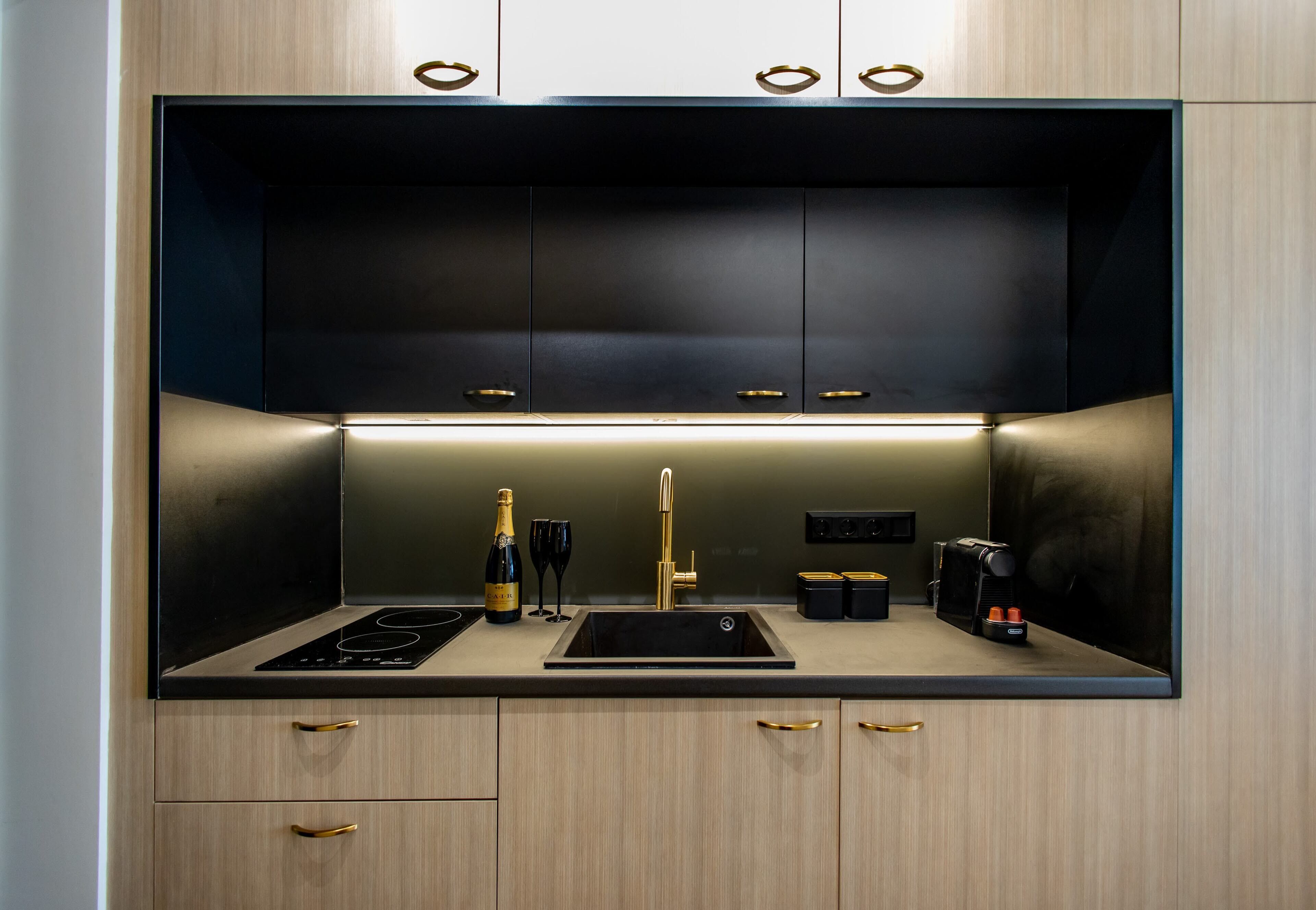 Cocina compacta con encimera negra, estufa integrada, lavamanos y cafetera. Hay dos cajones con tiradores dorados y un mueble pequeño sobre la encimera.