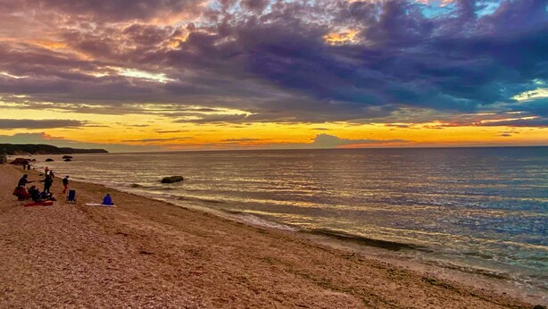 Een strand met mensen die zitten en staan in het zand, een rustige zee en een zonsonderganglucht met wolken.