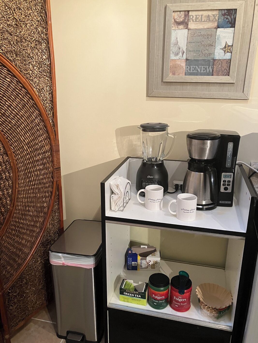 Une petite cuisine équipée d’une machine à café, d’un mixeur et d’une tasse posée sur le comptoir. On y trouve également une poubelle et une étagère avec des produits pour le thé et le café.