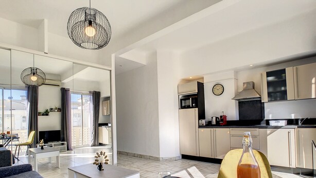 Un appartement moderne comprenant un salon, un coin repas et une cuisine. On y trouve une table à manger avec du pain, une bouteille de jus de fruits et un verre de vin rouge. La cuisine est équipée d’un four à micro-ondes, d’un grille-pain et d’une horloge murale.