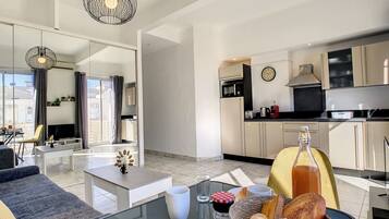 A modern apartment with a living room, dining area, and kitchen. There is a dining table with bread, a bottle of juice, and a glass of red wine. The kitchen has a microwave, toaster, and a clock on the wall.