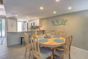 Uma sala de jantar com uma mesa redonda, cadeiras e uma cozinha ao fundo.