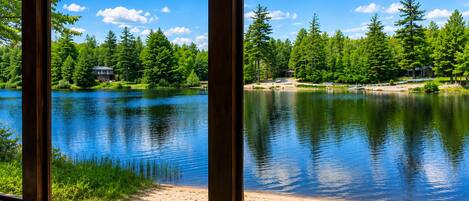 Blick auf einen See von innen, durch große Fenster, mit einem Sandstrand und bewaldeter Umgebung mit einer Holzhütte.