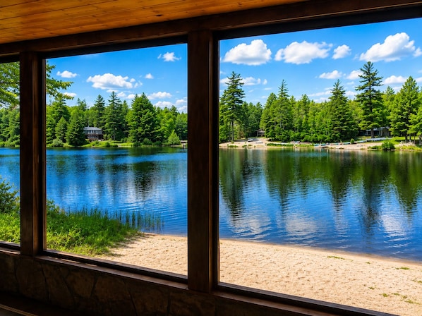 A lakeside view from inside a building with large windows, a sandy beach, and a forested area with a cabin.