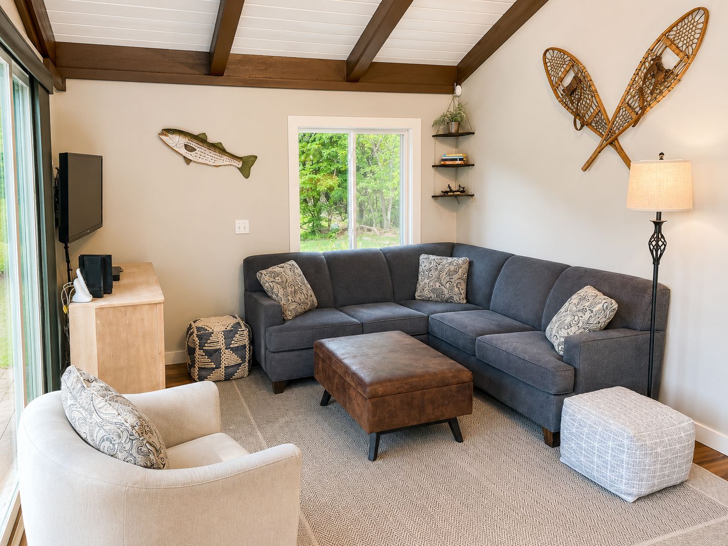 A living room with a gray sectional sofa, a white armchair, a brown ottoman, a patterned rug, a TV on a stand, and a window with a view of greenery.