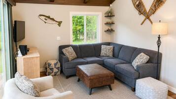 A living room with a gray sectional sofa, a white armchair, a brown ottoman, a patterned rug, a TV on a stand, and a window with a view of greenery.