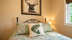 A bed with patterned bedding, a framed deer painting, and a lamp on the bedside table.