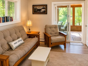 A cozy living room with a sofa, armchair, and a coffee table. There is a bookshelf with books, a lamp, and a framed picture on the wall.