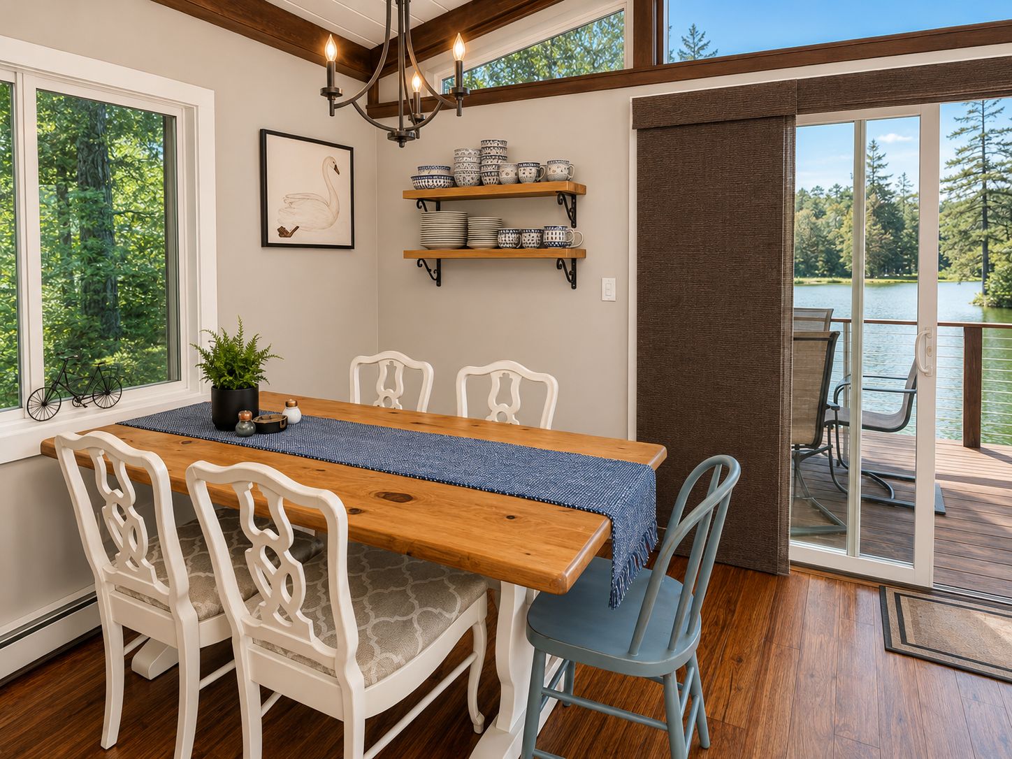 A dining area with a wooden table, white chairs, a blue table runner, and shelves with bowls and mugs.