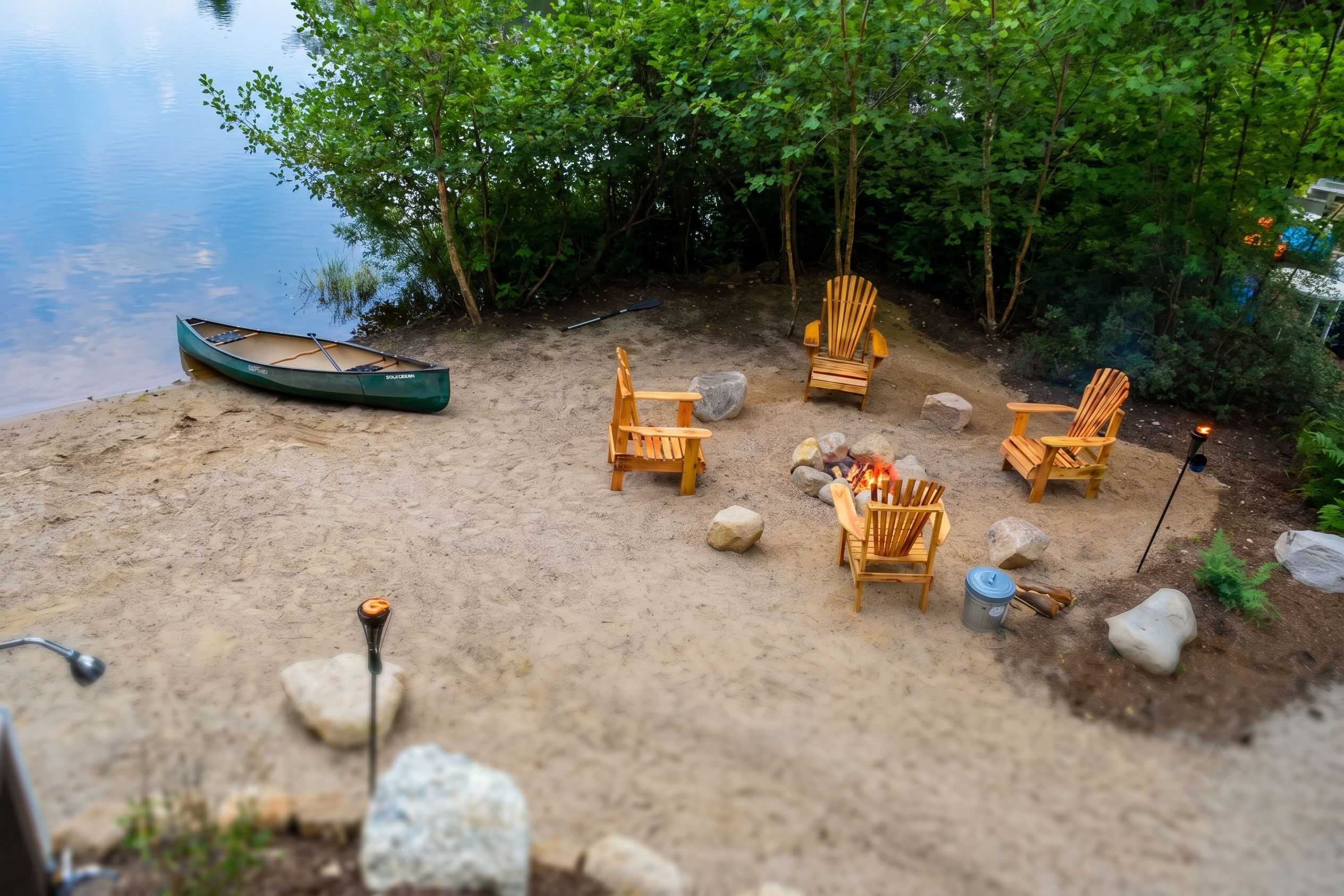 A sandy beach area with a canoe, wooden chairs, and a campfire.