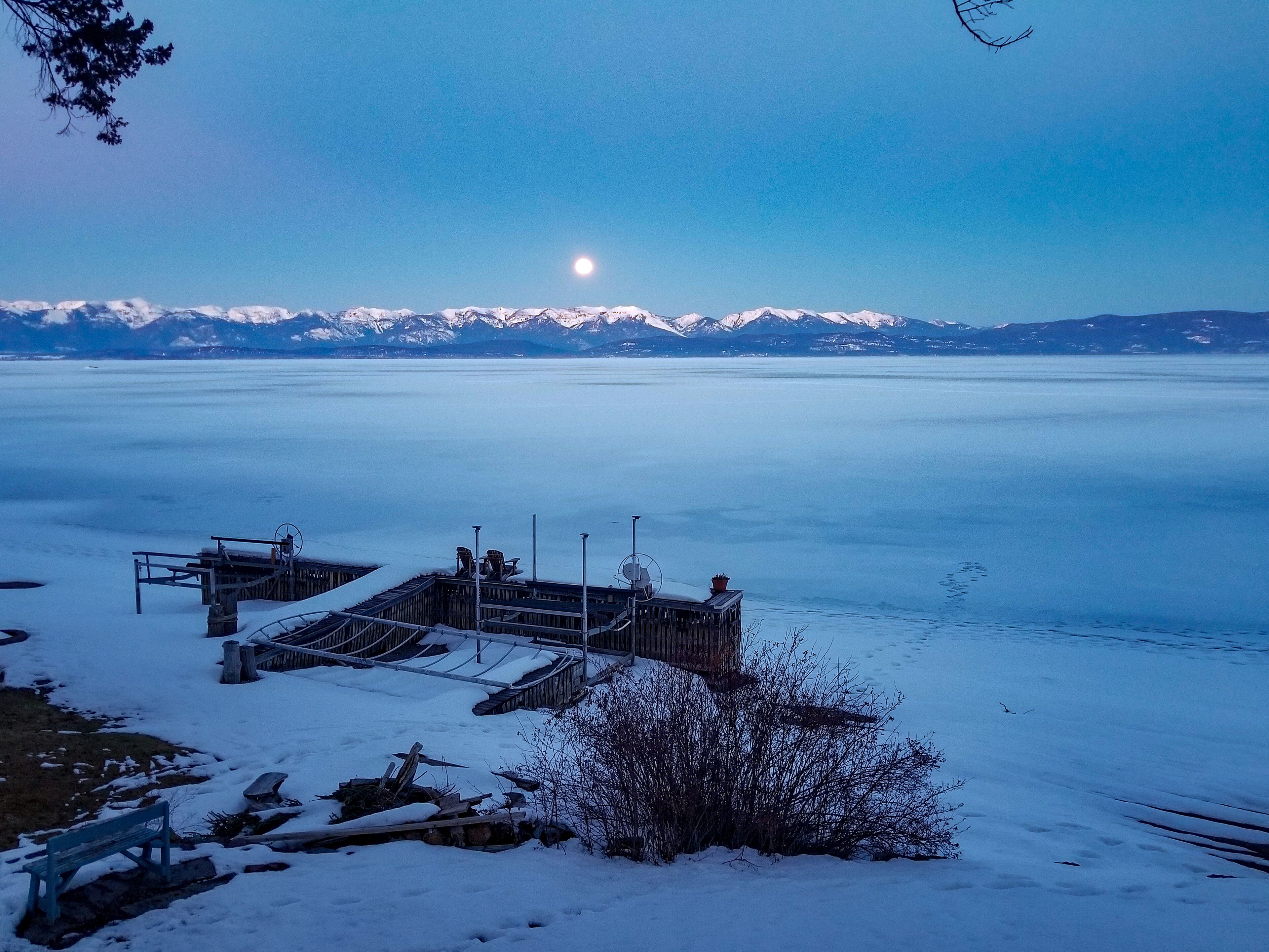 A snow-covered dock with a view of a calm lake and snow-capped mountains in the distance.