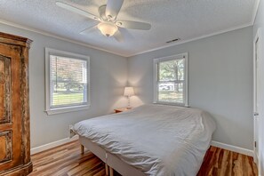 Une chambre à coucher avec un lit, deux fenêtres, un ventilateur de plafond et un plancher en bois.