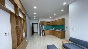 A modern kitchen with wooden cabinets, a built-in oven, and a sink. There is a blue bench and a wooden table in the living area. - Family Apartment with Seaview Balcony 03 (Nha Trang)