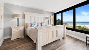 A bedroom with a large bed, two bedside tables, a nightstand, and a view of the beach through a large window.