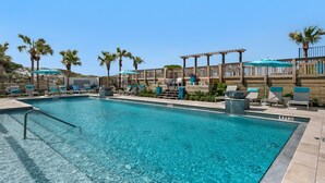 A clear blue pool with lounge chairs and umbrellas, surrounded by palm trees and wooden pergolas.