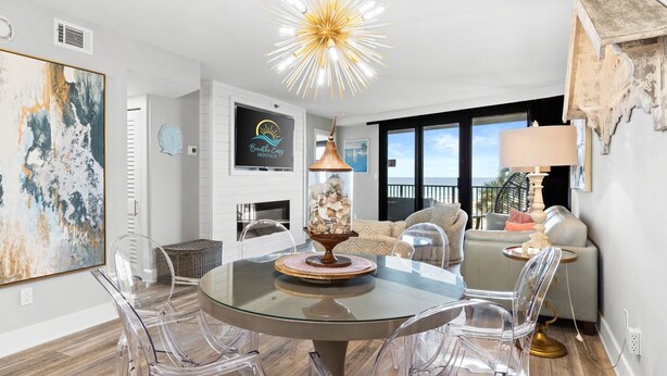 A modern living room with a glass dining table, clear chairs, a large abstract painting, a decorative lamp, and a view of the ocean through large windows.