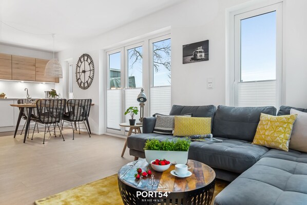 A modern living room with a grey sofa, a coffee table with a bowl of strawberries, and a dining area with a wooden table and chairs.