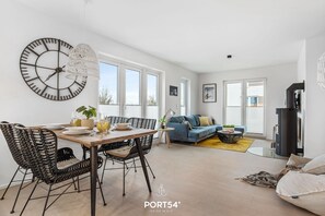 A modern living room with a dining area, featuring a large wall clock, a sofa, a fireplace, and a large window.