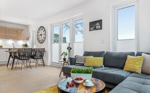 Una sala de estar moderna con un sofá gris, una mesa de centro con un bol de fresas, y un comedor con una mesa y sillas de madera.