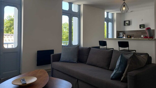 Una sala de estar moderna con un sofá, una mesa de centro y un televisor. Se ven ventanas amplias y una zona de cocina.