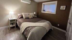 A bedroom with a bed, bedside tables, a window with lights, and a ceiling air conditioner. - Legal 2 bedroom suite conforms to the new 2024 Short Term Rental Regulations.  (Oliver)