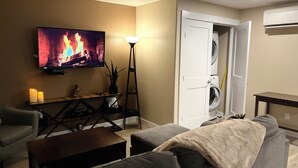 A living room with a television displaying a fireplace, a sofa, a coffee table, and a lamp. - Legal 2 bedroom suite conforms to the new 2024 Short Term Rental Regulations.  (Oliver)