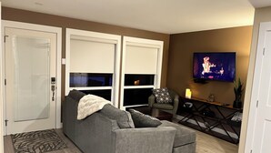 A living room with a grey sofa, a small armchair, a TV on the wall, and a wooden floor. - Legal 2 bedroom suite conforms to the new 2024 Short Term Rental Regulations.  (Oliver)