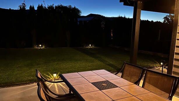 An outdoor patio with a tiled table and chairs, surrounded by a well-maintained lawn and a view of a distant mountain range. - Legal 2 bedroom suite conforms to the new 2024 Short Term Rental Regulations.  (Oliver)