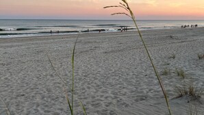 Een strand met zand en schaarse begroeiing, een heldere lucht en enkele mensen in de verte.