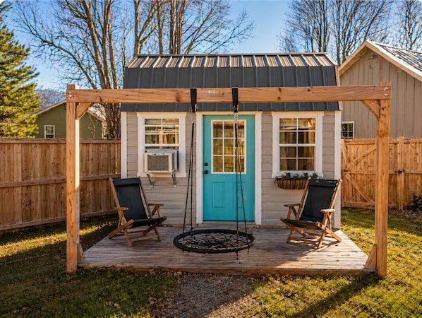 Une terrasse en bois avec deux chaises et une balançoire, une porte turquoise et un petit abri de jardin en arrière-plan.