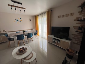 A modern living room with a dining area, a flat-screen TV, and a large window with curtains.