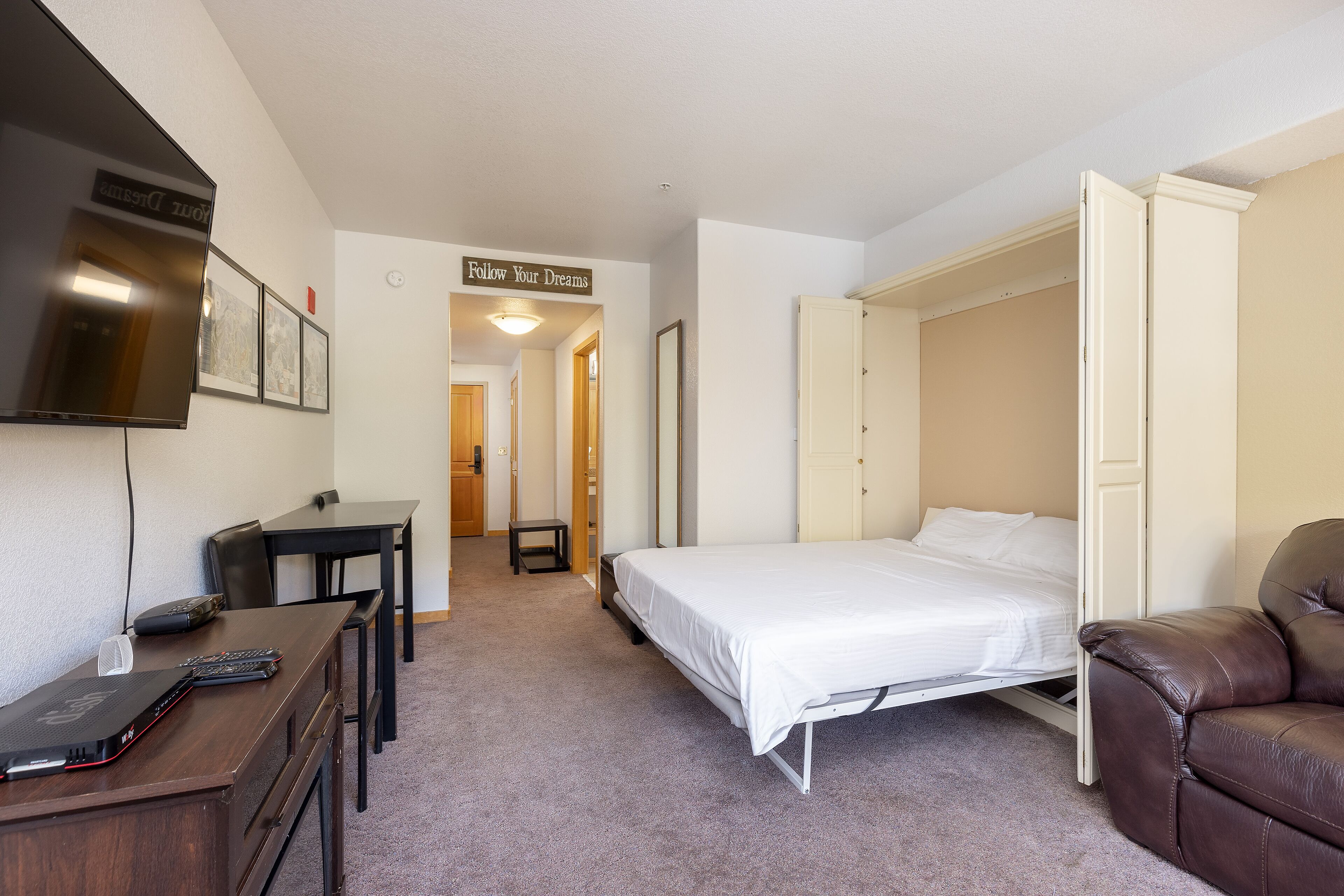A bedroom with a bed, a desk with a laptop, a TV, and a brown leather chair.