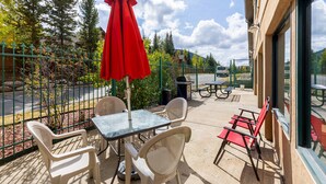 Un patio exterior con una mesa y sillas, una sombrilla roja, y vistas a árboles y una carretera.