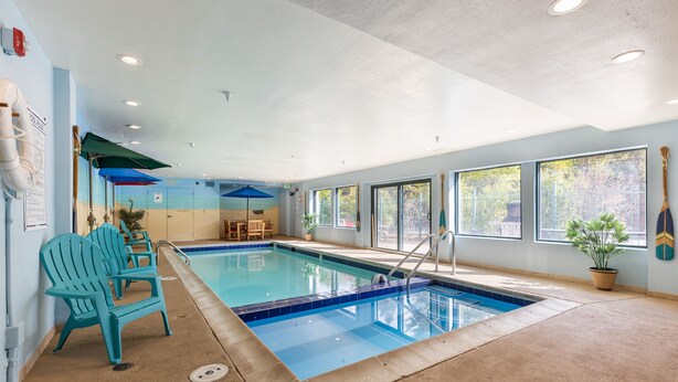 Piscina interior con agua azul cristalina, rodeada por un suelo color beige, y equipada con una silla de socorrista y una sombrilla azul.
