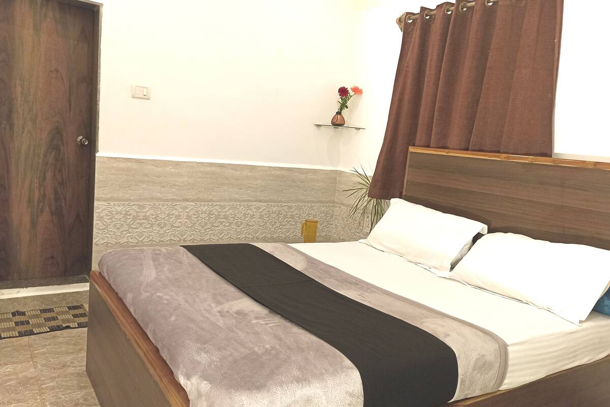 Una cama bien hecha con cabecera de madera, una mesita de noche con un florero y una cortina marrón.